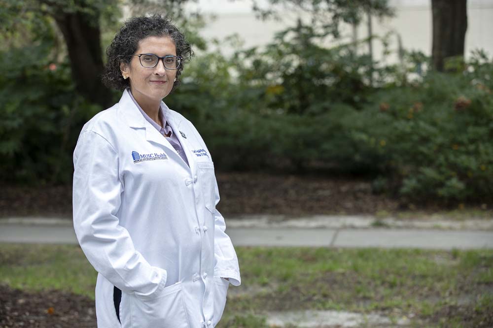 Woman wearing a white coat and glasses stands outside with greenery behind her.